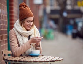 Female Customer Sending Text Message Sitting Outside Coffee Shop On Busy City High Street In Fall