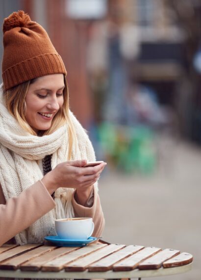 Female Customer Sending Text Message Sitting Outside Coffee Shop On Busy City High Street In Fall