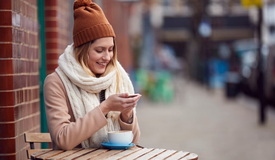 Female Customer Sending Text Message Sitting Outside Coffee Shop On Busy City High Street In Fall