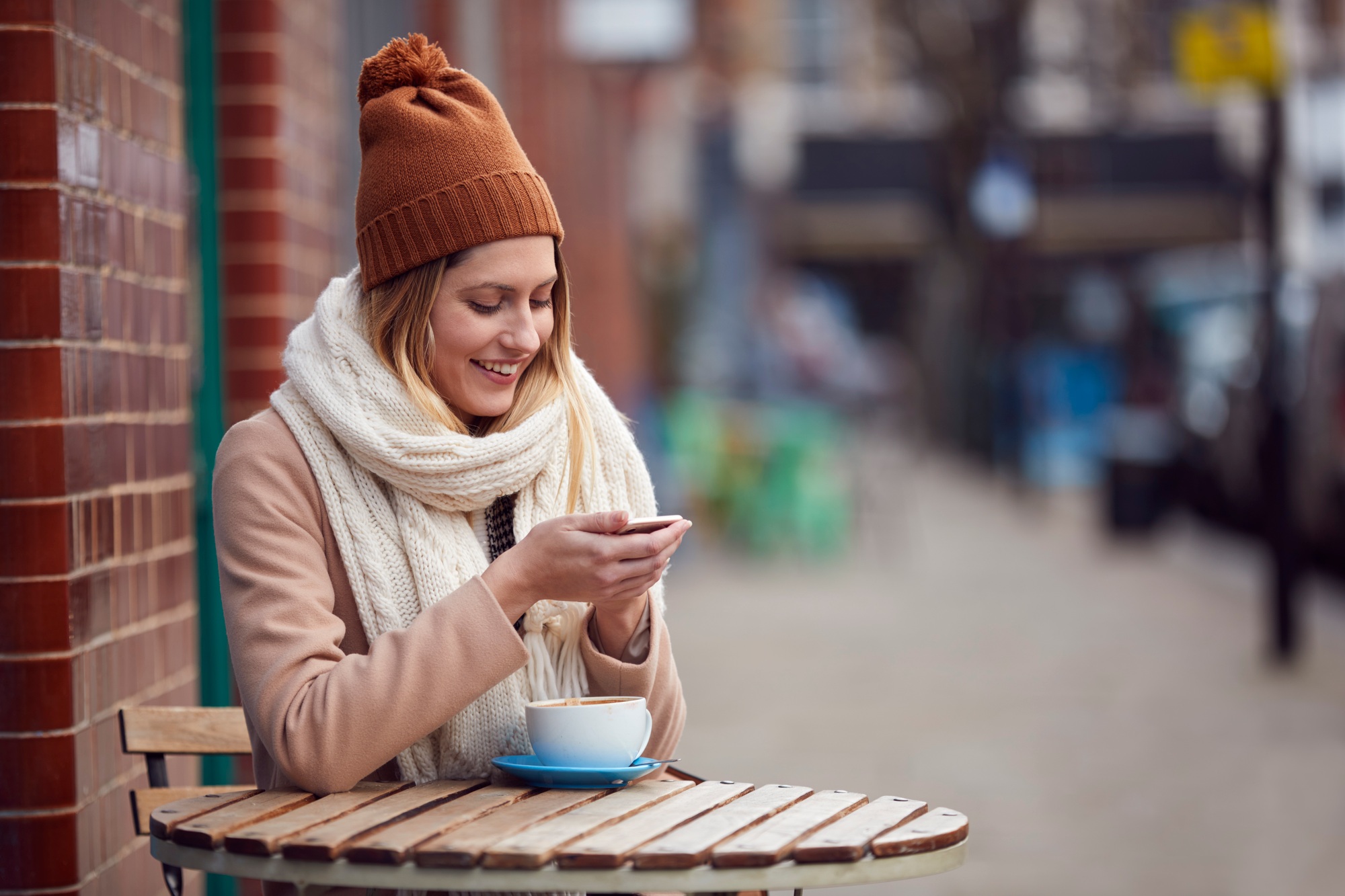 Female Customer Sending Text Message Sitting Outside Coffee Shop On Busy City High Street In Fall