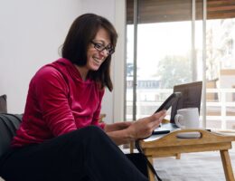 latin woman on vacation sitting in bed watching messages on her phone in morning while using laptop
