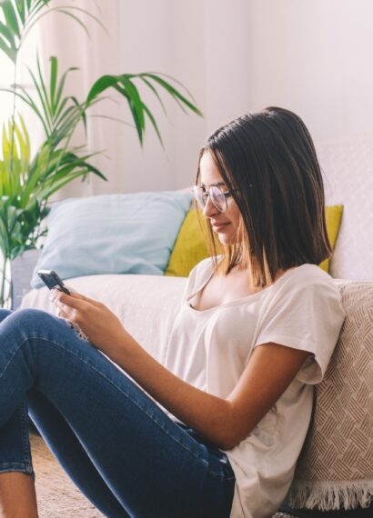 Serious woman in eyeglasses text messaging using mobile phone while sitting on floor