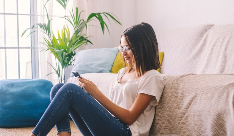 Serious woman in eyeglasses text messaging using mobile phone while sitting on floor