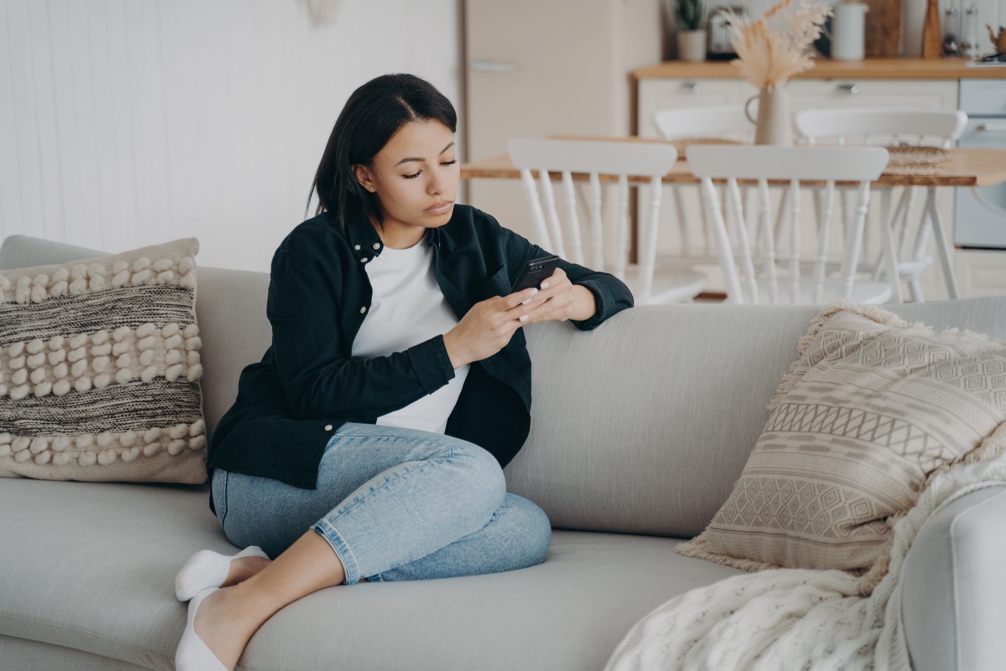 Young serious woman is texting on cellphone. Girl is sitting on on a couch and receiving messages.