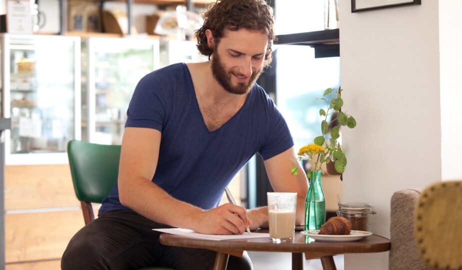 Smiling young man sitting in a coffee shop writing letter