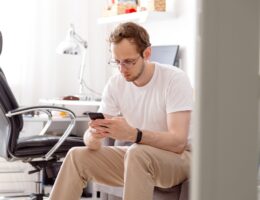 Young man in eyeglasses sitting on sofa sending text on mobile phone