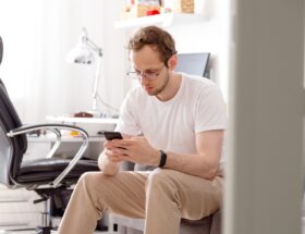 Young man in eyeglasses sitting on sofa sending text on mobile phone