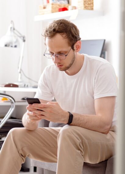 Young man in eyeglasses sitting on sofa sending text on mobile phone