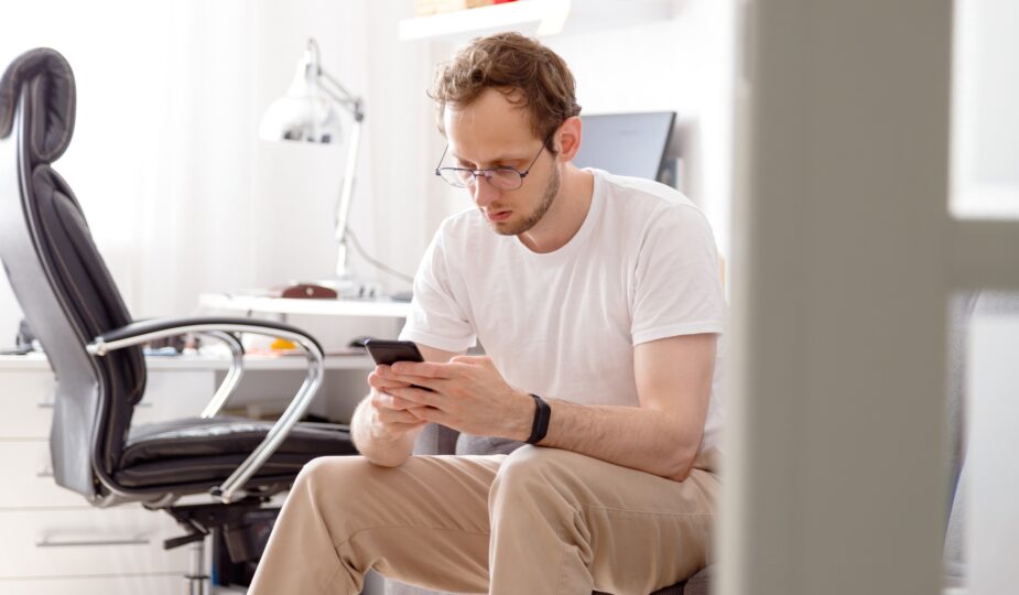 Young man in eyeglasses sitting on sofa sending text on mobile phone